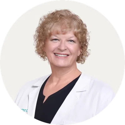Smiling middle-aged female medical professional with curly blonde hair wearing a white lab coat and black shirt.