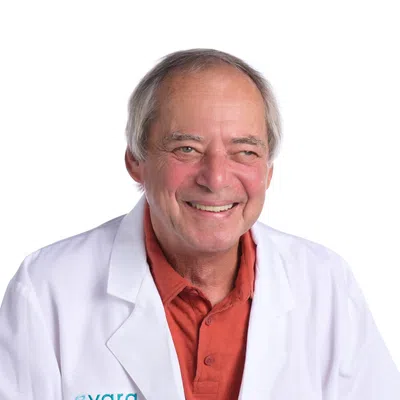 Smiling elderly male pharmacist wearing a white lab coat and burnt orange shirt against a white background