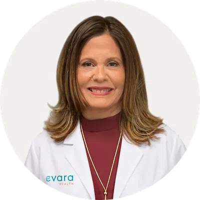 Smiling female healthcare professional wearing white coat and maroon top against a plain background.
