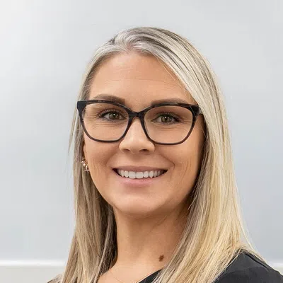 Smiling woman with long blonde hair wearing black glasses and a black top against a light gray background.