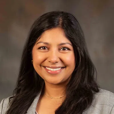 Smiling woman with long dark hair wearing a checked blazer and light blouse against a brown background