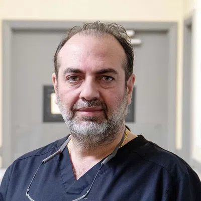 Middle-aged male healthcare professional with beard wearing navy scrubs and magnifying glasses around neck.
