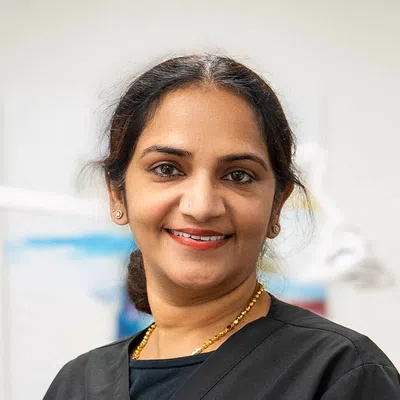 Smiling woman in a black medical scrub with gold necklace standing in a clinical setting.
