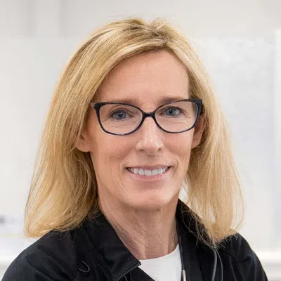 Smiling middle-aged woman with blonde hair and black glasses wearing a black jacket and white shirt.