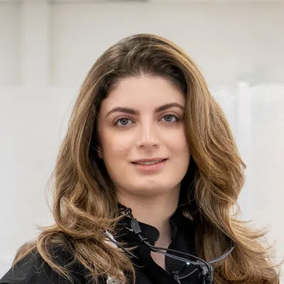 Female dentist with long hair wearing black coat and dental loupe around neck, smiling at camera.