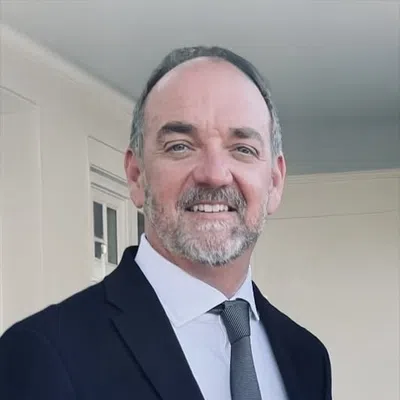 Smiling middle-aged man with salt-and-pepper beard wearing a black suit and gray tie indoors.