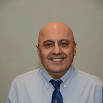Smiling bald man wearing glasses, light blue shirt, and blue tie with green stripes and yellow shield symbols.