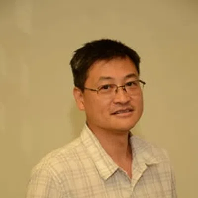 Man wearing glasses and a light plaid short-sleeve shirt standing against a beige background.