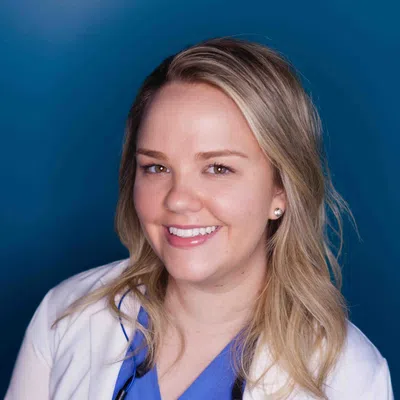 Smiling female healthcare professional wearing blue scrubs and white lab coat against a blue background