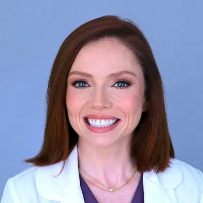 Smiling woman with red hair wearing a white coat and purple top against a light blue background.