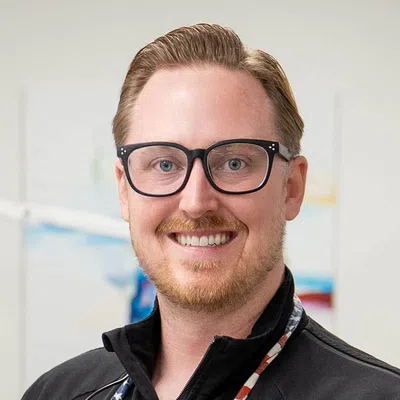 Smiling man with glasses and red beard wearing a black zip-up jacket against a light background.