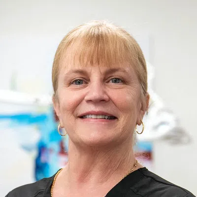 Smiling middle-aged woman with blonde hair wearing black scrubs and gold jewelry in a bright indoor setting