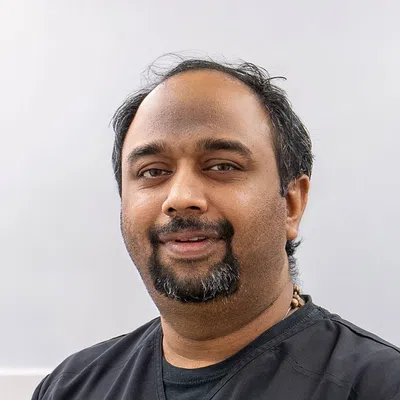 Smiling man with short black hair, beard, and black shirt against a plain gray background.