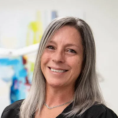 Smiling middle-aged woman with straight gray hair wearing a black top and silver necklace in a colorful background.