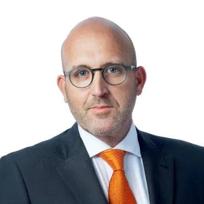 Bald man with glasses wearing a dark suit and bright orange tie against a white background