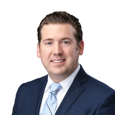 Professional man with brown hair wearing navy suit and light blue striped tie smiles against white background