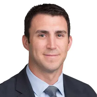 Professional man with short dark hair wearing a suit, light blue shirt, and patterned tie against white background