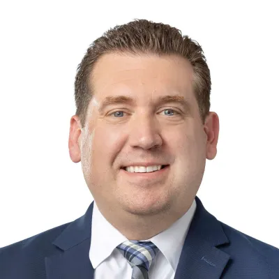 Professional headshot of a man in a dark suit, light blue shirt, and tie smiling against a white background
