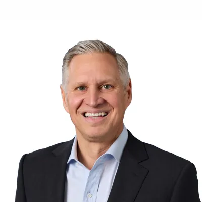 Close-up of a smiling man with light hair, blue eyes, wearing a gray blazer and white shirt against a white background.
