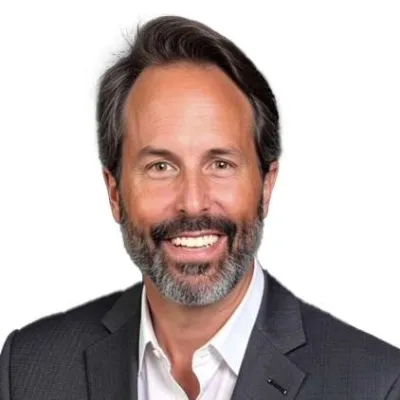 Smiling middle-aged man with salt and pepper beard wearing a dark suit and white shirt on white background