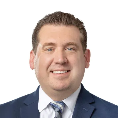 Professional headshot of a man in a dark suit, light blue shirt, and tie smiling against a white background