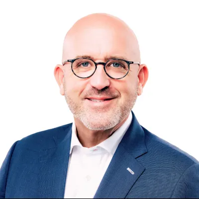 Bald man with glasses wearing a dark suit and bright orange tie against a white background