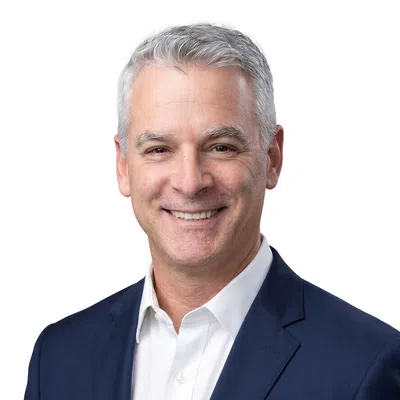 Smiling middle-aged man with gray hair wearing a navy suit jacket and white shirt on white background.