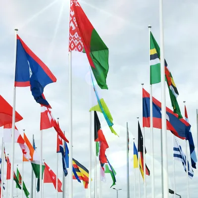 Multiple international flags waving on tall flagpoles against a cloudy sky background.