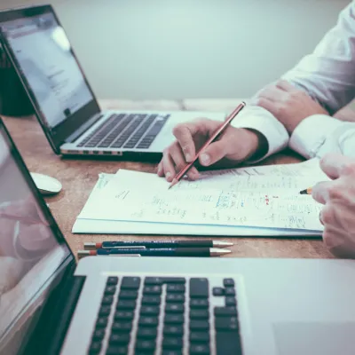 Two people working with laptops and documents on a wooden table, discussing and taking notes.