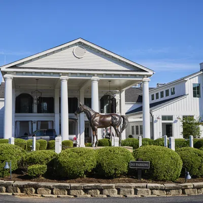 White colonial-style building with large columns, bronze horse statue, and manicured bushes under blue sky.