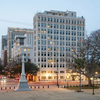 Cityscape at dusk featuring a historic monument, tall office buildings, and lit street lamps along the urban park.