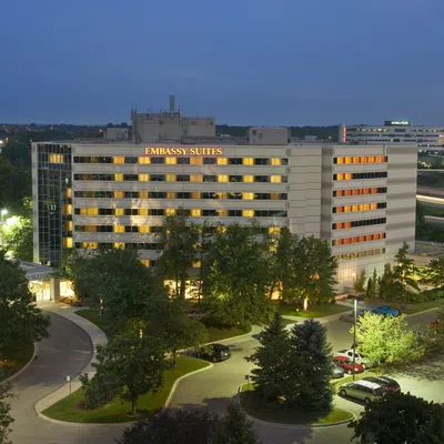 Night view of Embassy Suites hotel building with illuminated windows and surrounding trees in a parking area.