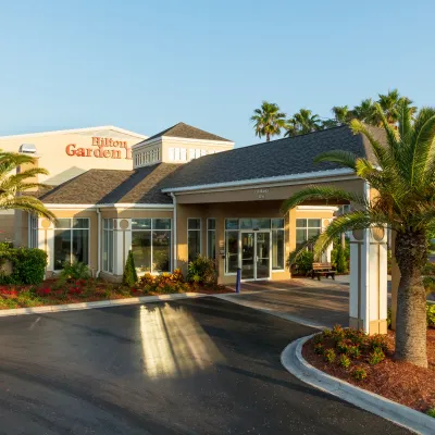 Exterior view of Hilton Garden Inn hotel entrance with palm trees and clear blue sky at sunset.