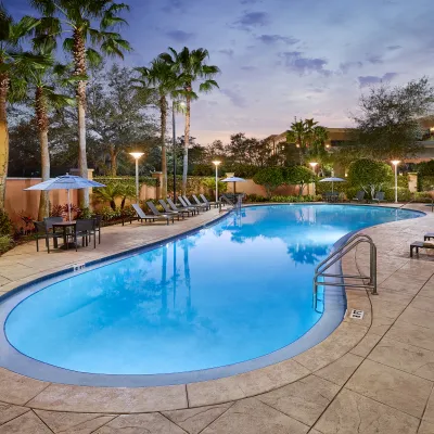 Evening view of an empty outdoor pool surrounded by palm trees and lounge chairs with soft lighting.