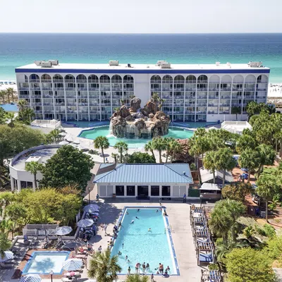 Oceanfront resort with large pool, rock waterfall feature, palm trees, and white sandy beach under clear blue sky.