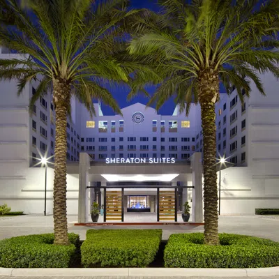 Sheraton Suites hotel entrance framed by palm trees and well-manicured bushes at dusk with lights on.