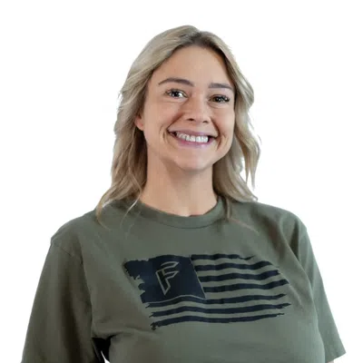 Smiling blonde woman wearing an olive green t-shirt with a black flag design and high-waisted blue jeans.