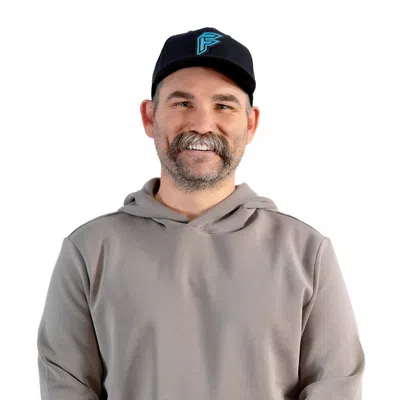 Smiling man with mustache wearing a gray hoodie and black cap standing with hands clasped in front on white background