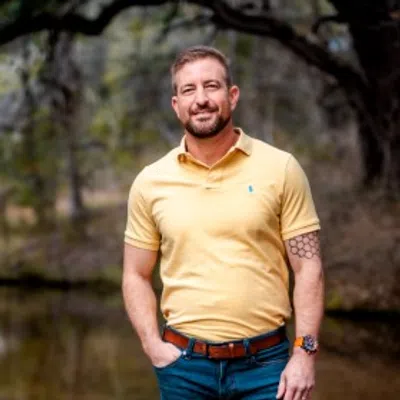 Smiling man with short hair and beard wearing yellow polo shirt and blue jeans outdoors with hands in pockets