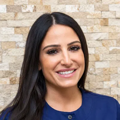 Smiling woman with long dark hair wearing a blue dental uniform against a stone wall background