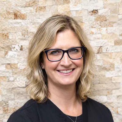 Smiling woman with blonde hair and glasses wearing black medical scrubs against a stone wall background