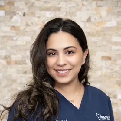 Portrait of a smiling female dentist in navy scrubs with long dark hair against a light stone background.