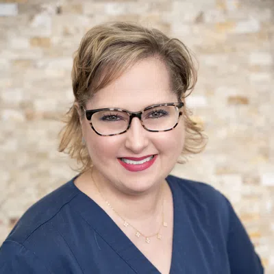 Smiling woman with short blonde hair and glasses wearing navy medical scrubs against a blurred brick wall.