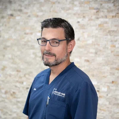 Male dental professional in blue scrubs with Midland Park Family Dentistry logo standing against stone wall.