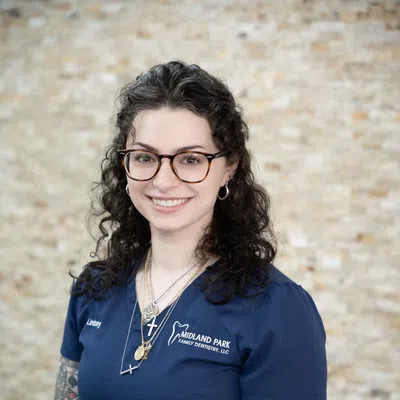 Smiling woman with glasses in blue Midland Park Family Dentistry uniform against brick wall background