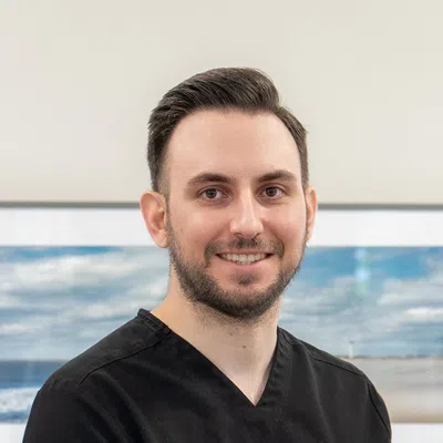 Smiling male dentist wearing black scrubs standing indoors with ocean-themed artwork in the background