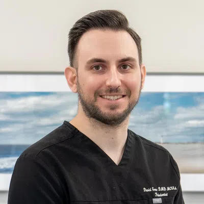 Smiling male dentist wearing black scrubs standing indoors with ocean-themed artwork in the background