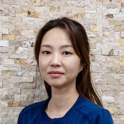 Portrait of a confident Asian woman with long hair wearing a blue jacket against a textured stone wall.