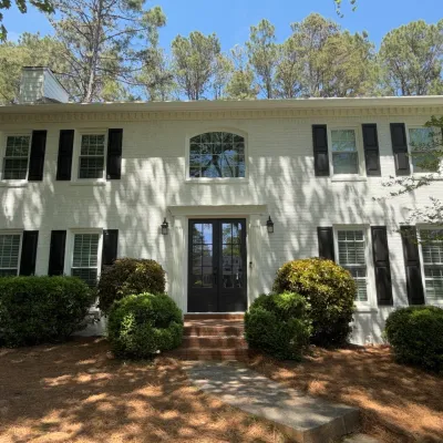 White two-story house with black shutters, glass front door, surrounded by green bushes and tall pine trees.