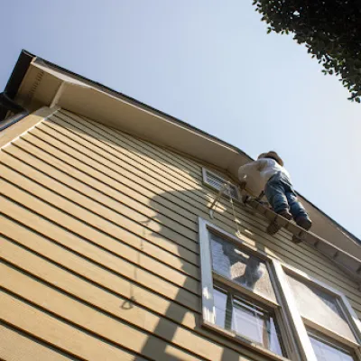 Person wearing protective gear painting the upper exterior of a yellow wooden house on a sunny day.